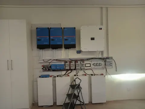 Three blue inverters and a white grid-tie inverter above a wired DC and AC board, with four battery cabinets below