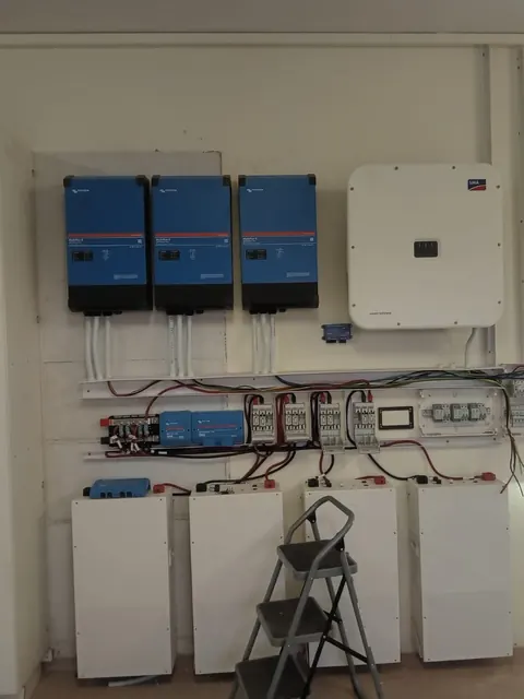 Three blue inverters and a white grid-tie inverter above a wired DC and AC board, with four battery cabinets below