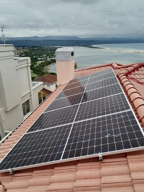 Rows of solar panels fitted to a terracotta tile roof in Knysna, with the lagoon, the beach and the mountains behind