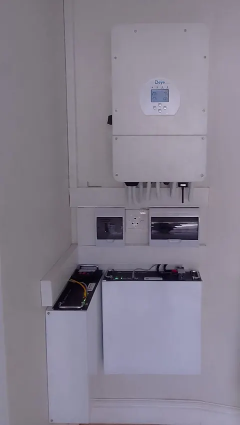 A white hybrid inverter above two distribution boards and a boxed battery, fitted neatly into a wall nook
