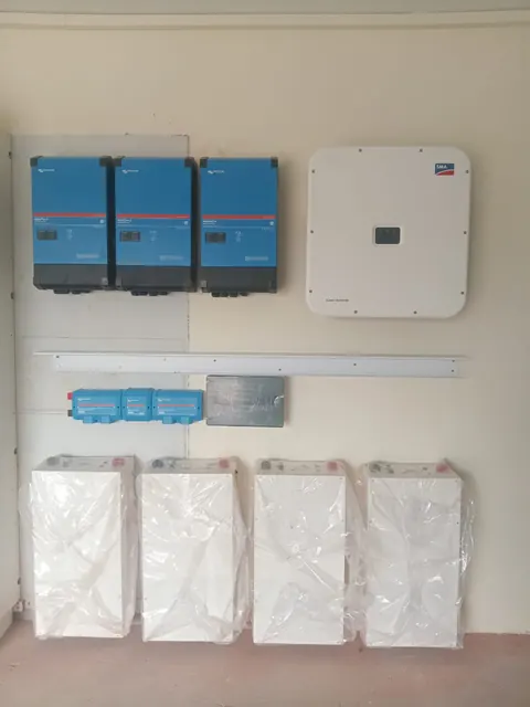 Three blue inverters and a white inverter mounted on a wall, with four new batteries still in their wrapping below
