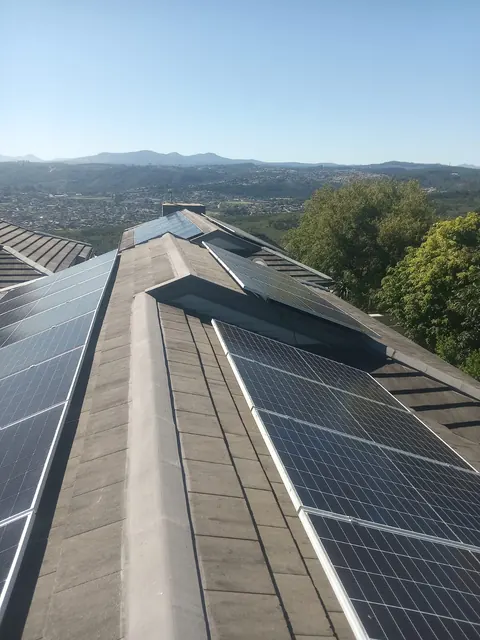 Solar panels on both sides of a grey tile roof ridge, with trees and the hills of the town behind