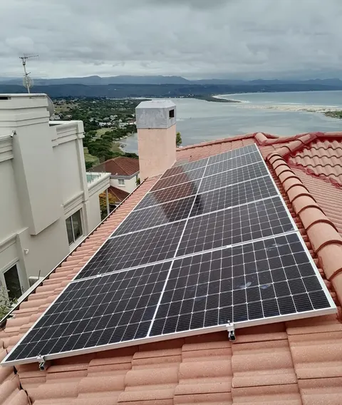 Rows of solar panels fitted to a terracotta tile roof in Knysna, with the lagoon, the beach and the mountains behind