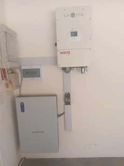 A white Sunsynk inverter on a wall above a small DB and a grey battery cabinet